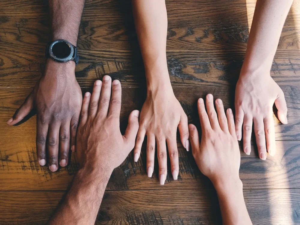 Hands of different skin tones on a wooden surface.
