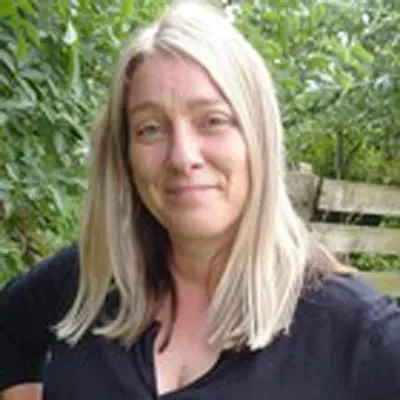 A woman with blonde hair smiles, wearing a black top, with a green background.