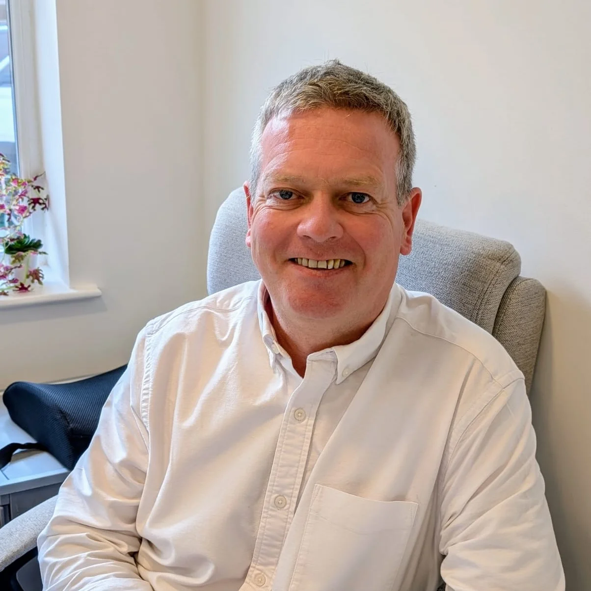 Man wearing a white shirt smiles. He sits in a chair with a window behind him.