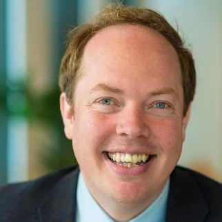 Close-up of a smiling man with light skin and blonde hair. He wears a suit jacket.