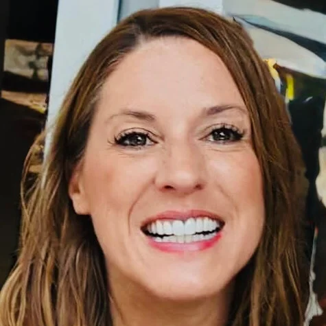 Close-up portrait of a smiling woman with long, wavy brown hair and dark eyes. She is wearing light makeup and has a bright white smile.