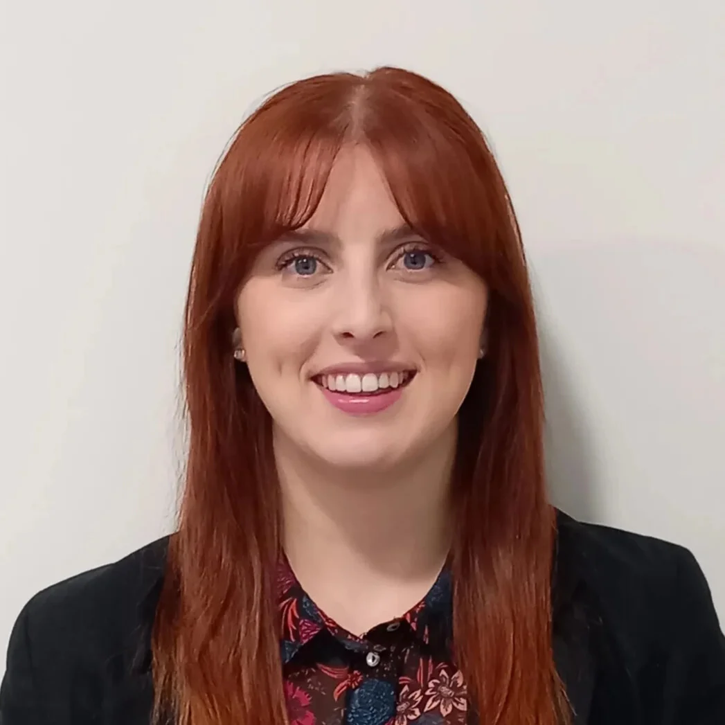 A young woman with long red hair and blue eyes smiles, wearing a black blazer over a floral shirt.