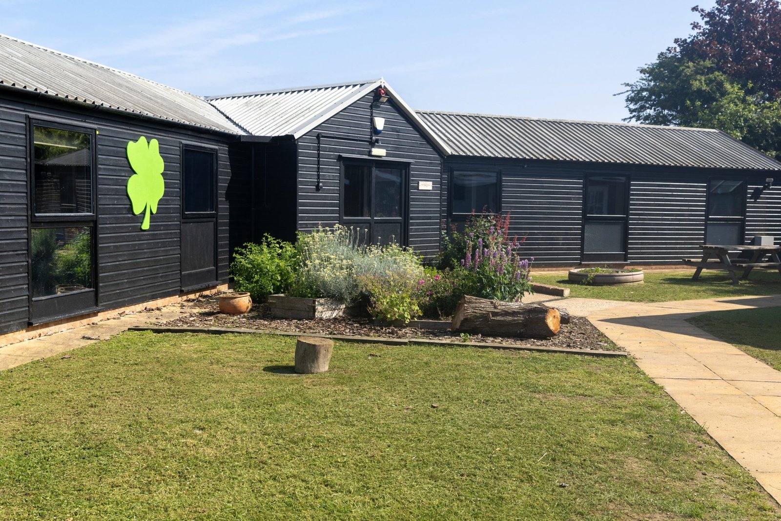 Black wooden stables with a green shamrock on the wall, surrounded by a grassy yard, flower beds, and a paved path.